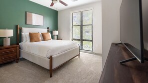 Un dormitorio con cama, mesitas de noche, ventilador de techo, ventana con persianas y televisor de pantalla plana sobre un soporte de madera.
