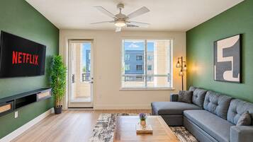 A living room with a flat-screen TV, a ceiling fan, a sofa, a coffee table, and a large window with blinds.