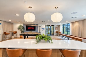 Un área de lounge moderna con mesa de billar, asientos cómodos, una pantalla grande mostrando 'NETFLIX' y un ventanal con vistas.