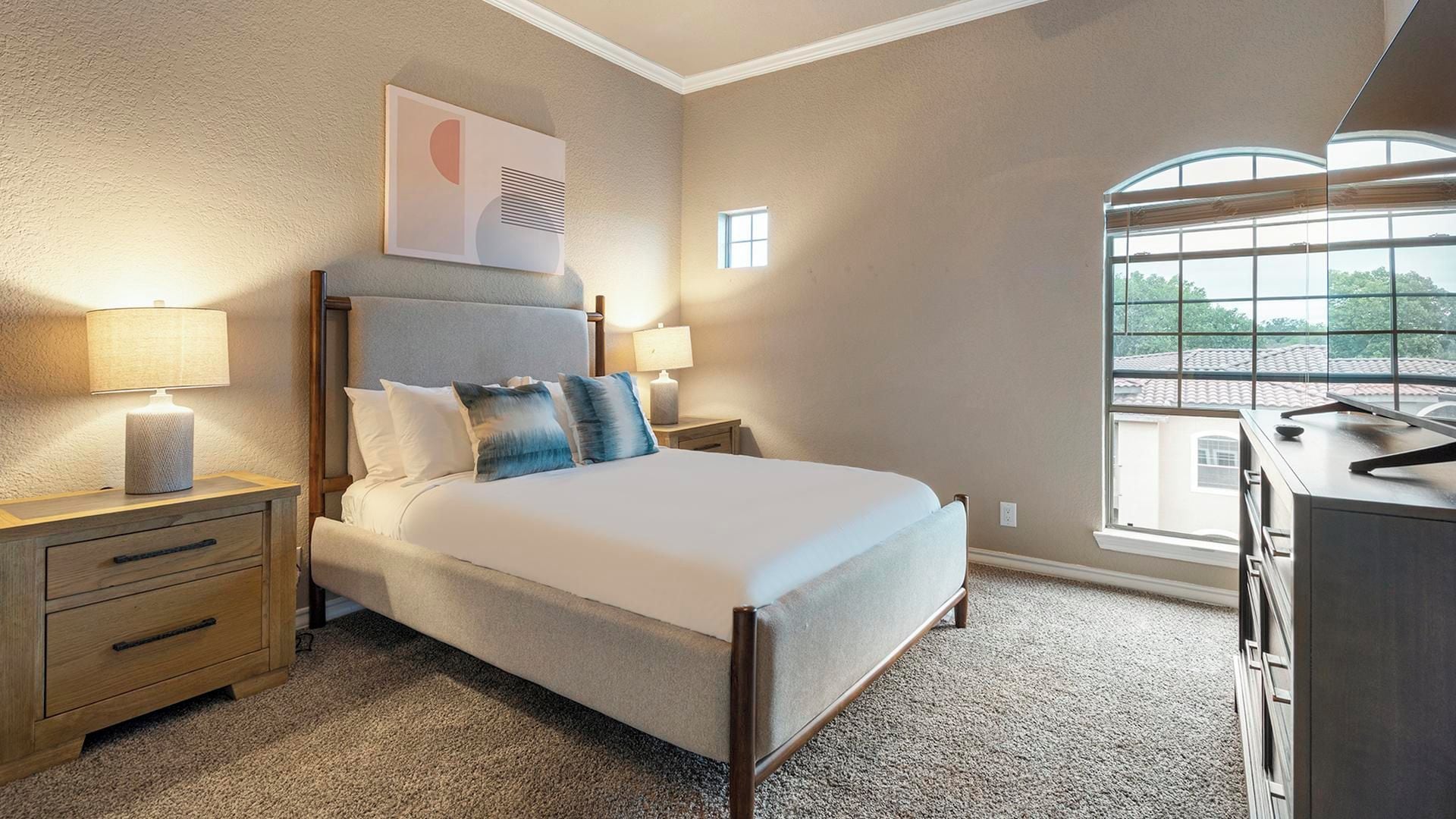 A hotel room with a bed, two bedside tables, a desk, a window, and a wall-mounted air conditioning unit.