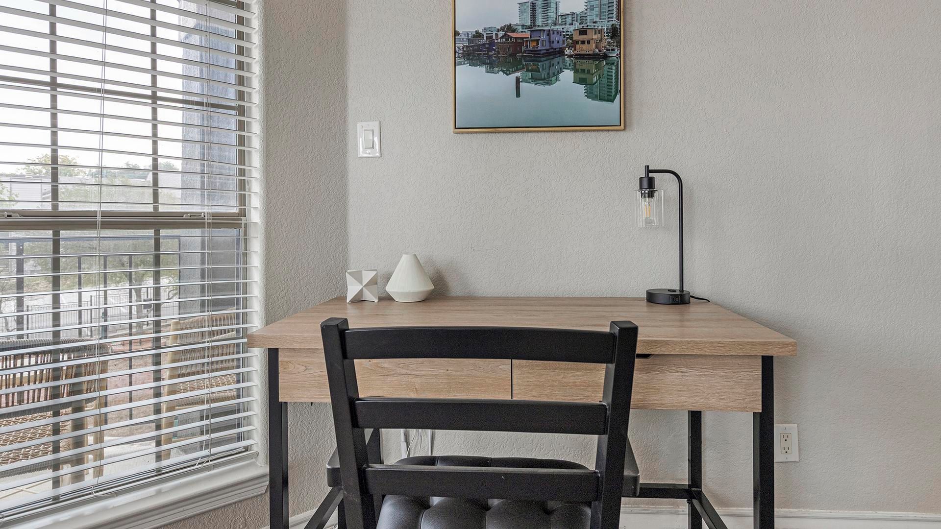 Una habitación con un escritorio de madera, una silla negra, una lámpara de pie y un cuadro de un paisaje urbano en la pared.