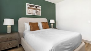 A neatly made bed with white bedding and two brown pillows, flanked by two bedside tables with lamps, against a green wall with a framed picture above. - Landing | Modern 1BD in Whitestone (Fort Worth)