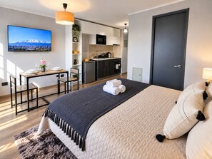 A modern hotel room with a bed, a desk, a kitchenette, and a TV displaying a cityscape. - Estudio Metro Toesca y Club Hípico (Santiago)