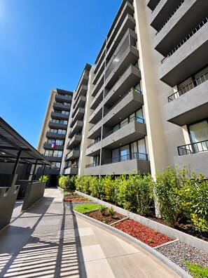 A modern multi-story building with balconies, a covered walkway, and landscaped areas with plants. - Estudio Metro Toesca y Club Hípico (Santiago)