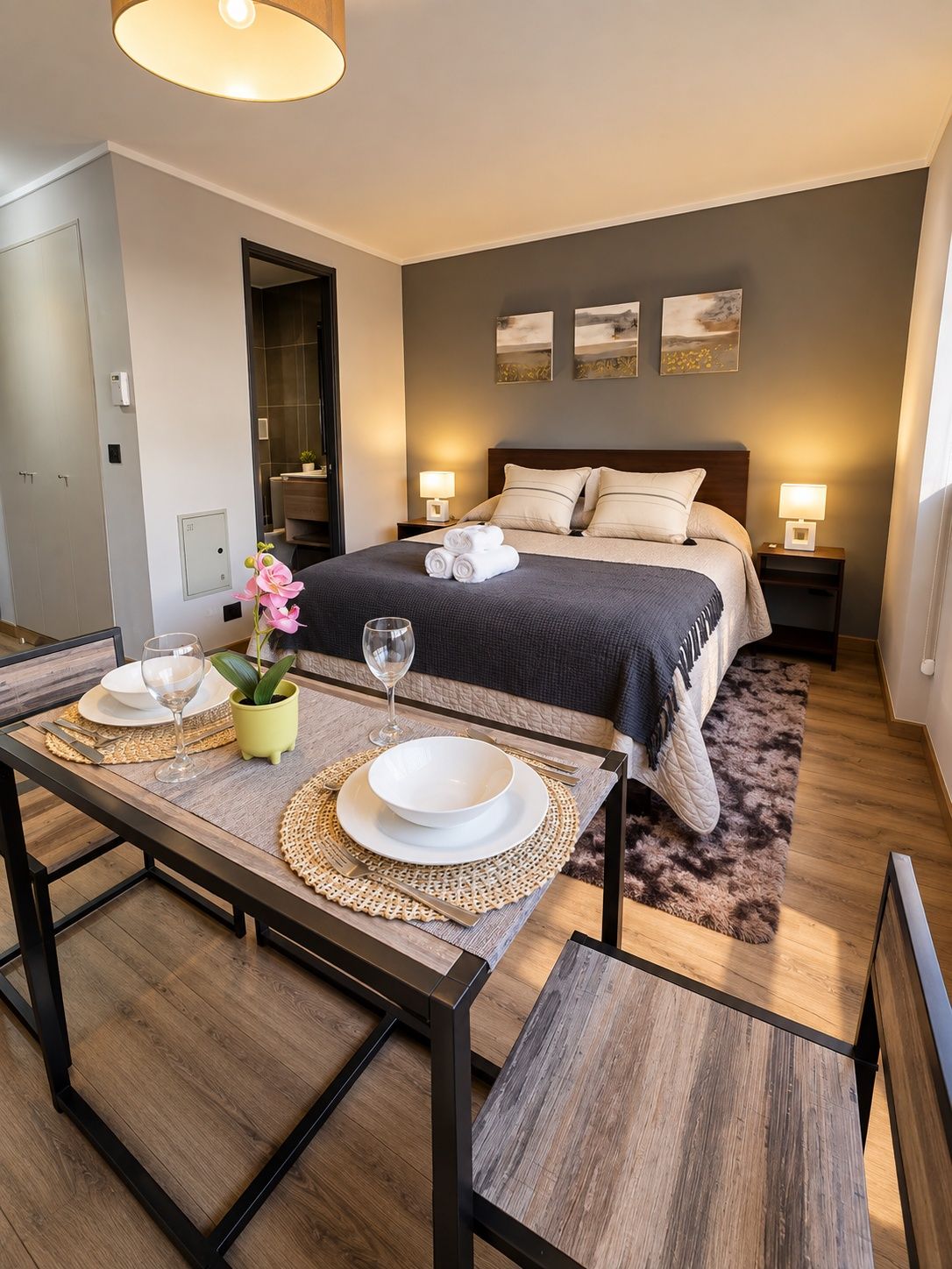 Une chambre d’hôtel moderne avec un lit soigneusement fait, une table à manger dressée pour deux personnes et une petite plante en pot.