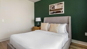Ein ordentlich bezogenes Bett mit weißer Bettwäsche, einer Nachttischlampe und gemusterten Kissen vor einer grünen Wand.