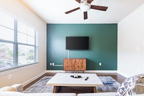 Ein Zimmer mit Deckenventilator, einem an der Wand montierten Fernseher, einem Holzschrank, einem Tisch und einem Fenster mit Jalousien.