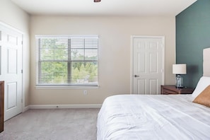 A bedroom with a large window, a door, a bedside table with a lamp, and a bed with white bedding. - Landing | Chic 3BD, Gym, Pool (Oxford)