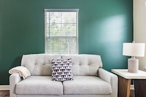 A room with a green wall, a light-colored sofa with a patterned pillow, a window with blinds, and a lamp on a side table. - Landing | Chic 3BD, Gym, Pool (Oxford)