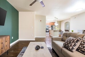 A living room with a sofa, coffee table, and a TV. There is a kitchen in the background with modern appliances. - Landing | Incredible 3BD, Pool, Gym (Oxford)