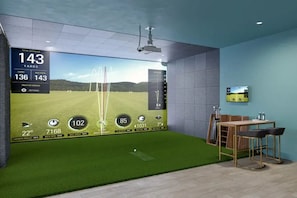 Et værelse med et stort skærm der viser golf-simulation, et lille bord med stole, og et fjernsyn på væggen.