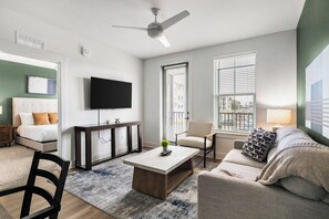 A modern living room with a flat-screen TV, a sofa, a coffee table, and a chair. There is a bed with a headboard in the background. - Landing | Amazing 2BD, Gym (Pensacola)