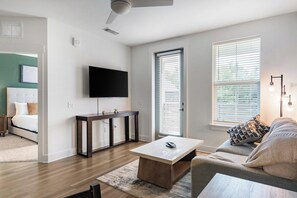 A modern living room with a flat-screen TV mounted on the wall, a wooden coffee table, a sofa, and a large window with blinds. - Landing | Modern 1BD, Gym (Pensacola)