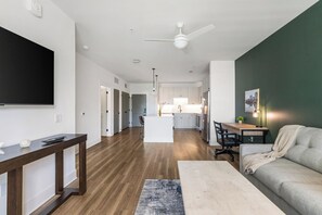 A modern living room with a sofa, a coffee table, and a TV. There is a kitchen in the background with a dining area. - Landing | Beautiful 1BD, Gym (Pensacola)