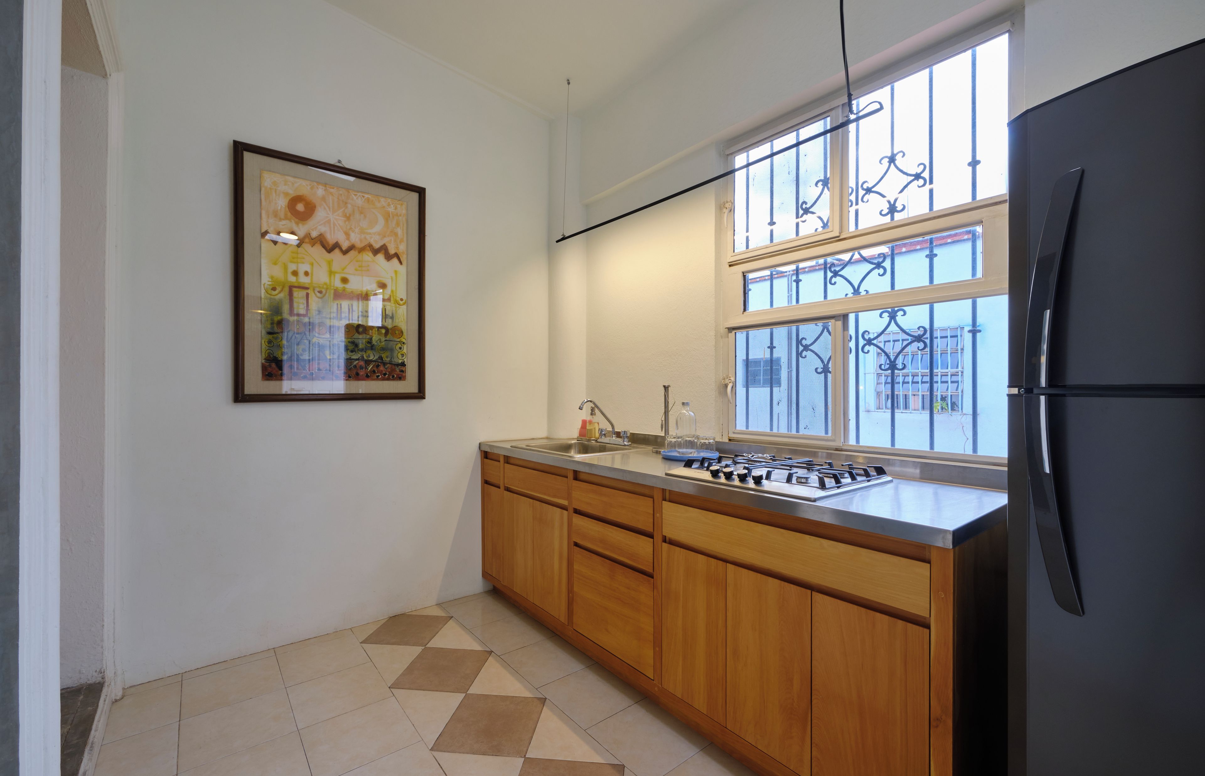 A kitchen with a black refrigerator, wooden cabinets, a tiled floor, a window with metal bars, and a framed picture on the wall.