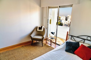 Quarto com cama, cadeira, mesa pequena e vista para prédios pela janela.