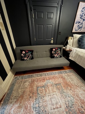 A room with a dark door, a grey sofa with floral cushions, a patterned rug, and a bed with a blue pillow. - Comfortable, Super Clean & Close to NYC (Hoboken)