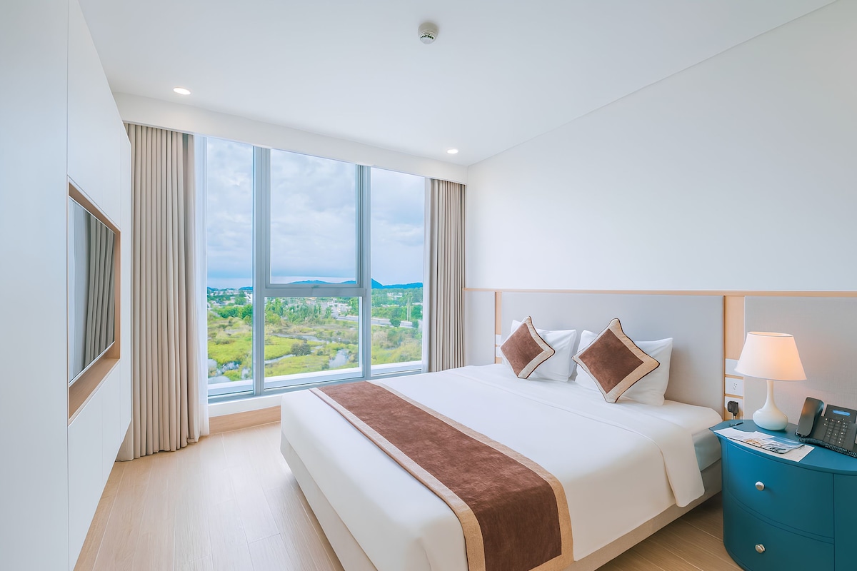 Ein Hotelzimmer mit einem großen Bett, einem Nachttisch, einer Lampe und Blick auf eine Landschaft durch das Fenster.