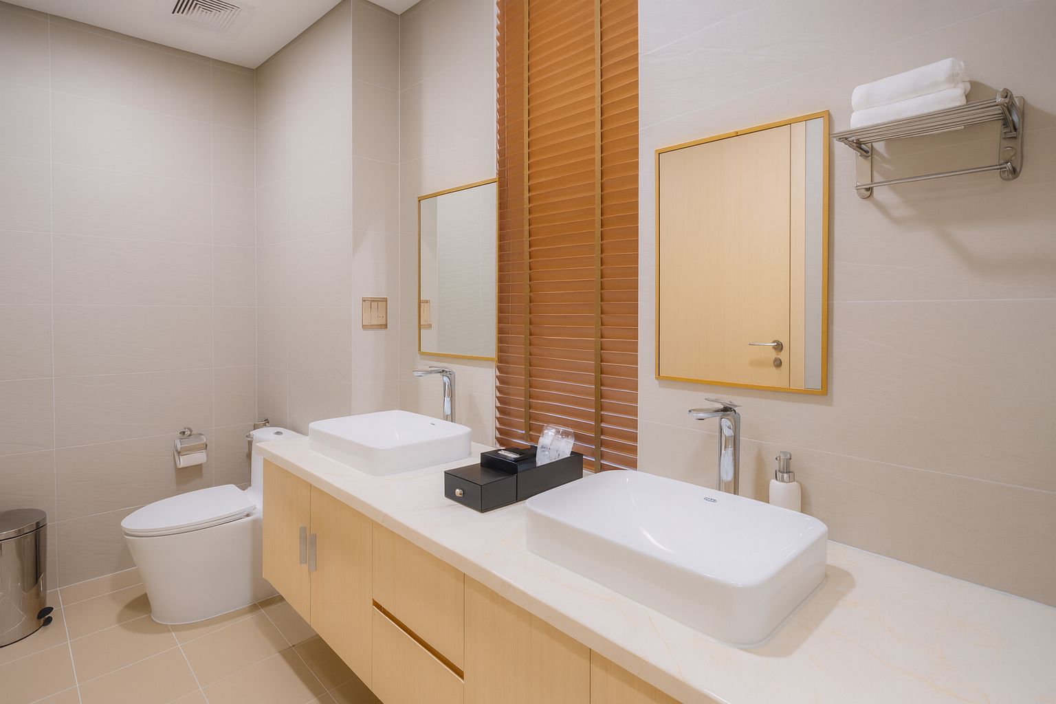 A modern bathroom with two sinks, a toilet, and a trash bin.