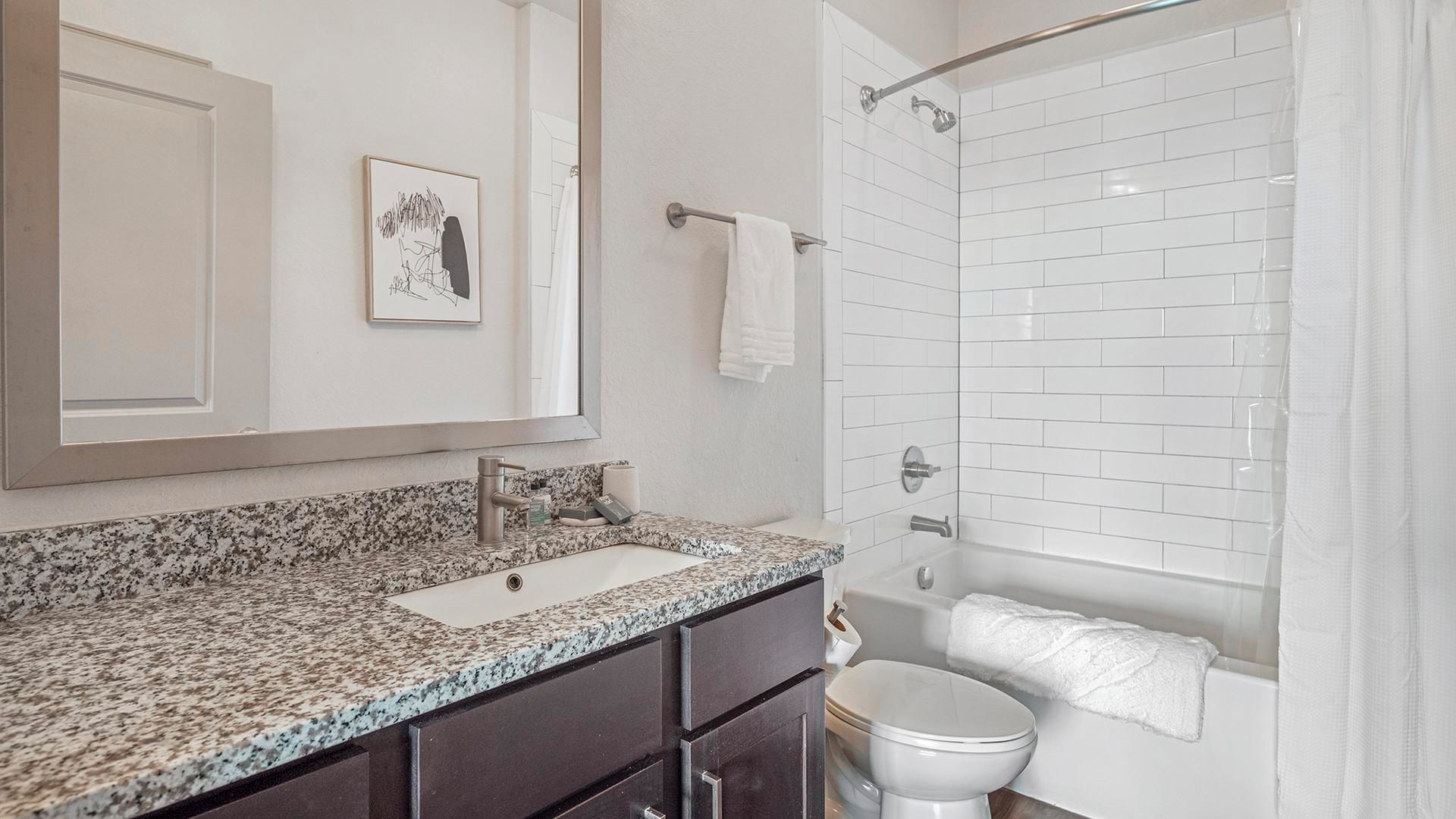 Un baño con encimera de granito, un lavabo blanco, inodoro y ducha con puerta de vidrio.