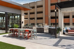 An outdoor patio area with a barbecue grill, seating, and a view of a parking garage. - Landing | Modern 1BD, Pool, Yoga Studio (Green Bay)