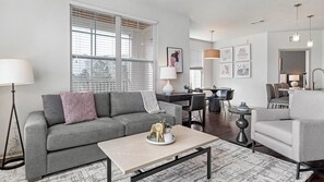 A modern living room with a grey sofa, a coffee table, and a dining area with a table and chairs. - Landing | Incredible 1BD, Yoga Studio, Gym (Green Bay)