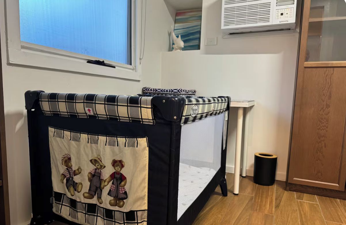 A crib with a plaid cover and a teddy bear design, positioned in a room with a window, a wall-mounted air conditioner, and a wooden cabinet.