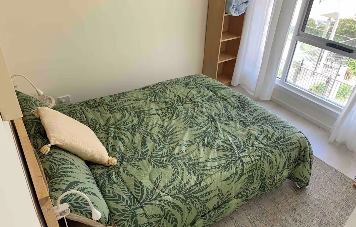 A single bed with a leaf-patterned bedspread and a pillow, a bedside table with a lamp, and a window with a view of the outside.