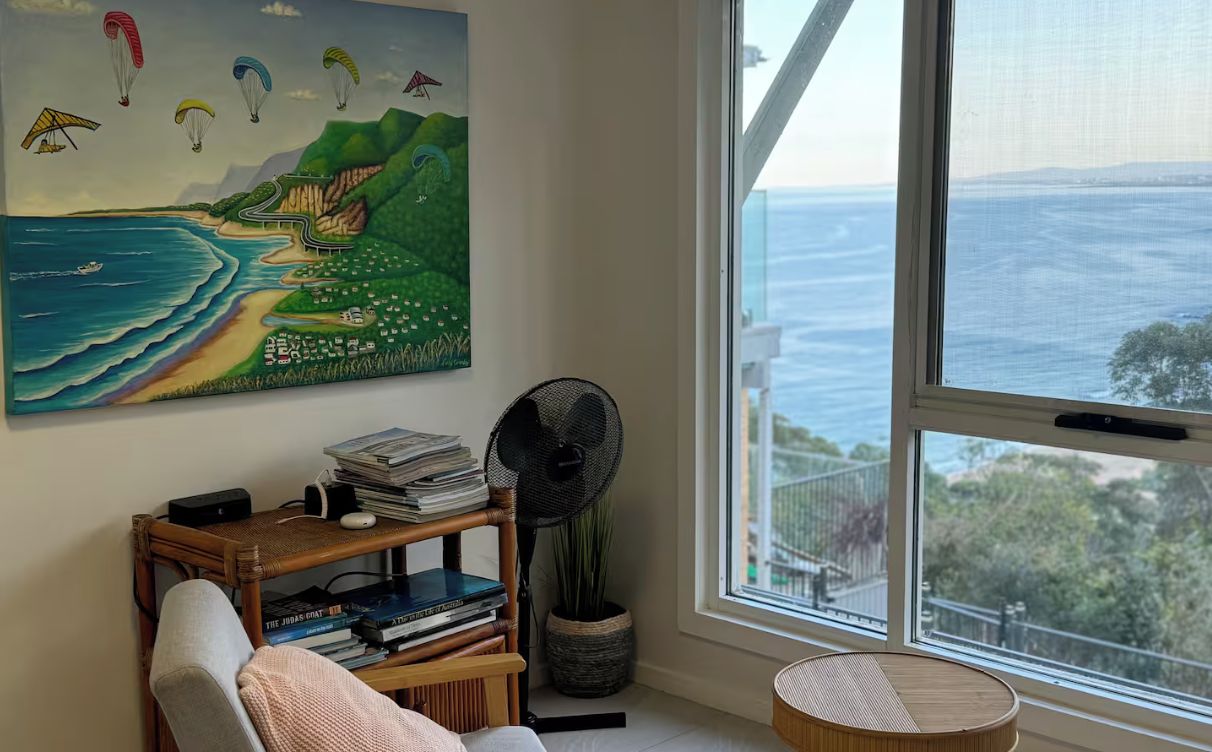 Une pièce dotée d’une grande fenêtre donnant sur un paysage côtier, d’un tableau représentant des parapentistes, d’une bibliothèque et d’un ventilateur.