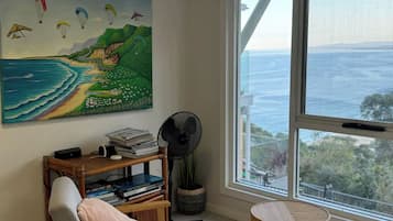 Une pièce dotée d’une grande fenêtre donnant sur un paysage côtier, d’un tableau représentant des parapentistes, d’une bibliothèque et d’un ventilateur.