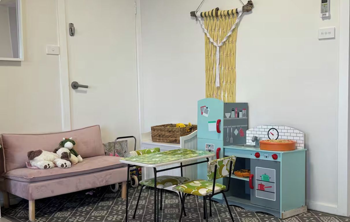 Une pièce avec un canapé, une petite table, des chaises, une cuisine miniature pour enfants et un rideau.