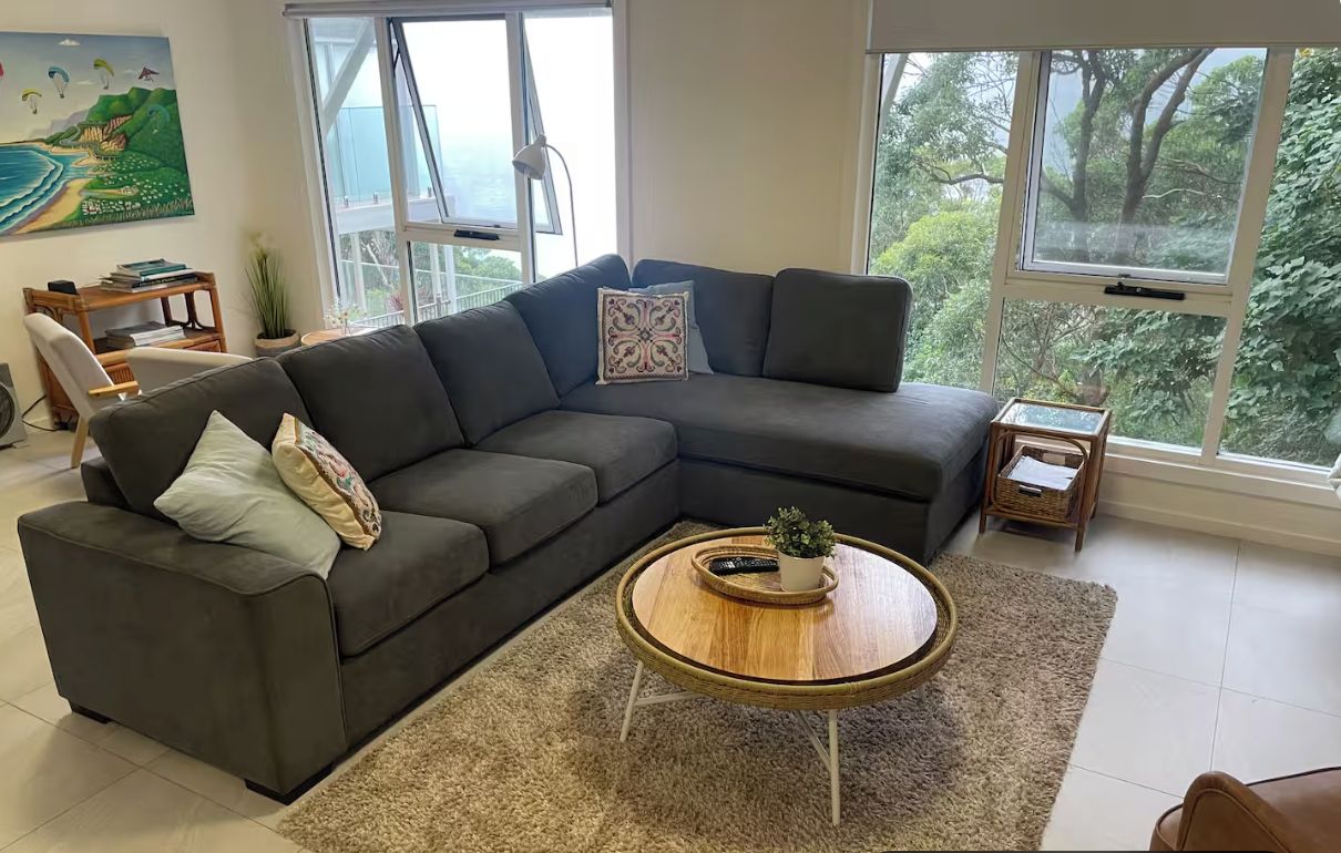 Ein modernes Wohnzimmer mit einem Ecksofa, einem runden Couchtisch und großen Fenstern mit Blick ins Freie.