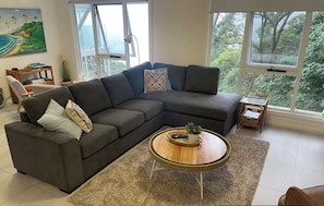 Ein modernes Wohnzimmer mit einem Ecksofa, einem runden Couchtisch und großen Fenstern mit Blick ins Freie.