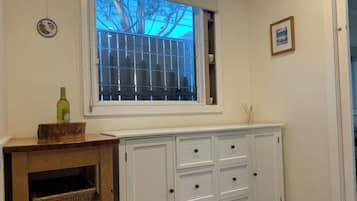 Une pièce avec une fenêtre, une armoire blanche avec des tiroirs, une petite table sur laquelle se trouve une bouteille, et un panier.