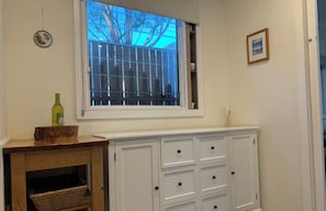 Ein Zimmer mit Fenster, einem weißen Schrank mit Schubladen, einem kleinen Tisch mit einer Flasche und einem Korb.