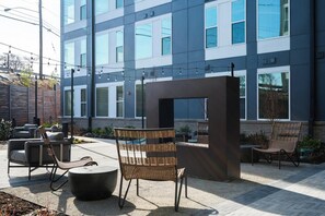 An outdoor seating area with wicker chairs and a round table, a modern building with large windows in the background, and a clear sky. - Landing | Chic 2BD, Rooftop Lounge (Richmond)