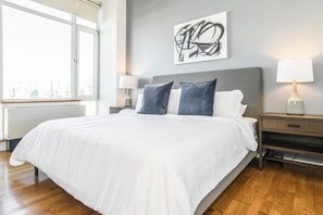 A modern bedroom with a large bed, a grey headboard, two bedside tables with lamps, and a cityscape view through a large window. - Landing | Incredible 2BD, Pool, Gym (Roy)