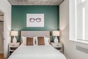A bedroom with a bed, two bedside tables, a headboard, a wall-mounted picture of glasses, and a window. - Landing | Stylish 2BD, Gym, Pool (Roy)