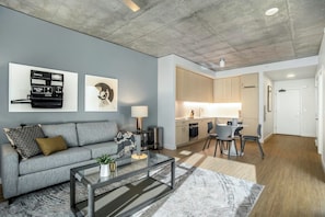 A modern living room with a grey sofa, a glass coffee table, and a kitchen area with wooden cabinets and a dining table. - Landing | Stylish 2BD, Gym, Pool (Roy)