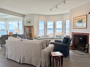 Un salon spacieux doté d’une grande fenêtre donnant sur une plage, d’une cheminée et d’un coin repas.