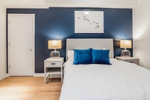 Ein Schlafzimmer mit einem Bett, zwei Nachttischen mit Lampen, einer Tür und einer Wand mit einem gerahmten Bild.