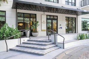 A hotel entrance with a modern design, featuring a set of stairs leading to the door, a large planter with greenery, and a metal railing. - Landing | Incredible 1BD, Pool (Tampa)