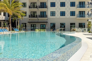 A swimming pool with lounge chairs, a palm tree, and a multi-story building with balconies in the background. - Landing | Incredible 1BD, Pool (Tampa)