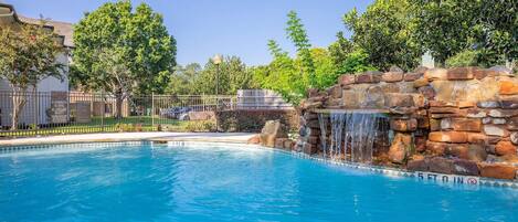A swimming pool with a waterfall feature, surrounded by a fence and trees.