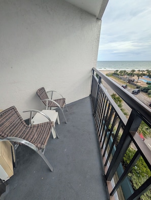 A balcony with wicker chairs and a small table, overlooking a beach with a pool and palm trees. - Oceanview-Corner Condo-2 Private Balconies-Hot Tubs, Heated Pools, Lazy Rivers. (Myrtle Beach)