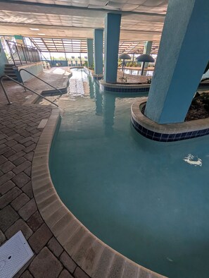 Una piscina interior con bordes azulejados, barandillas de metal y una zona cubierta con sillas y sombrillas.