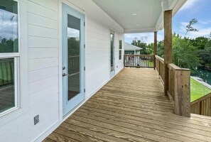 A covered wooden deck with a railing, a glass door, and a view of a green landscape. - Bay St. Louis Coastal Getaway. 3BR/3BA Near Old Town (Bay St. Louis)