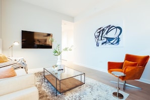 Ein modernes Wohnzimmer mit einer Couch, einem Glastisch, einer Pflanze, einem Gemälde und einem leuchtend orangefarbenen Sessel.