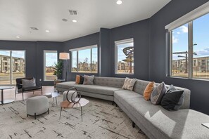 A modern living room with a grey sectional sofa, a round coffee table, and a grey armchair. Large windows offer views of the outside. - Landing | Modern 2BD, Game Room, Clubhouse (Littleton)