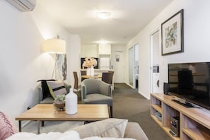 A hotel room with a sofa, a dining table, a TV, and a kitchenette in the background. - Stylish 2 Bedroom in Southbank (Southbank)